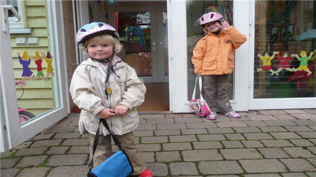 Two young girls wearing bicycle helmets and carrying school bags standing outside nursery doors