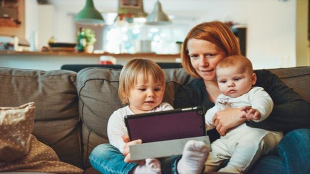 babies and woman sitting on sofa while holding baby and watching on tablet