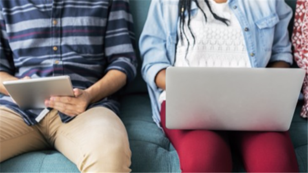 Man and woman sitting side by side on a sofa. He is looking at a tablet and she is working on a laptop.